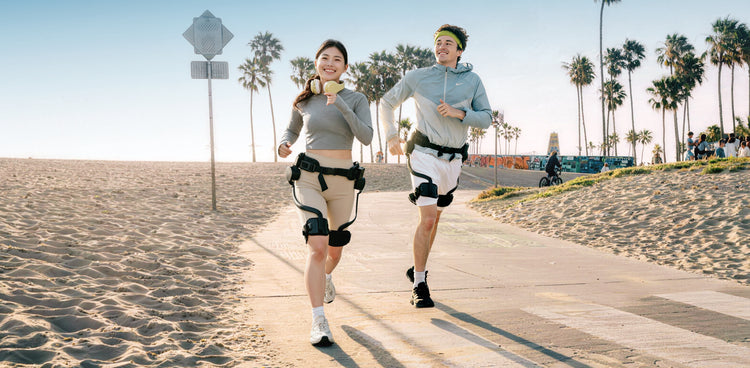 A man and a woman running on a sunny beach boardwalk wearing the Ascentiz exoskeleton, with palm trees and a sandy beach in the background.