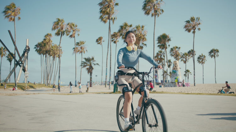 A cyclist wearing an Ascentiz hip exoskeleton rides a conventional bicycle along a seaside path.