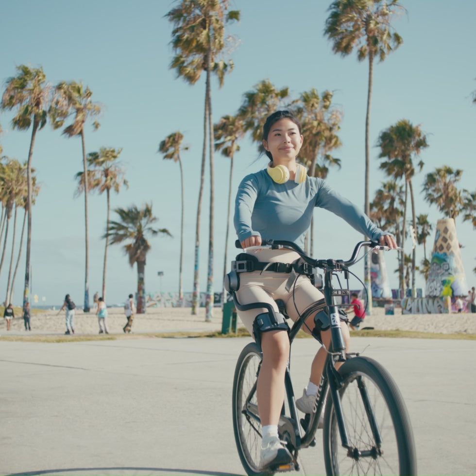 A cyclist wearing an Ascentiz hip exoskeleton rides a conventional bicycle along a seaside path.