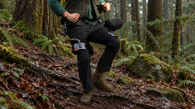 A hiker wearing an Ascentiz hip exoskeleton steps along a damp woodland trail after light rain.