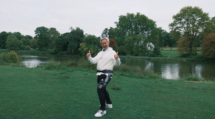 A person wearing an Ascentiz hip exoskeleton walks beside a lake and gives two thumbs up.