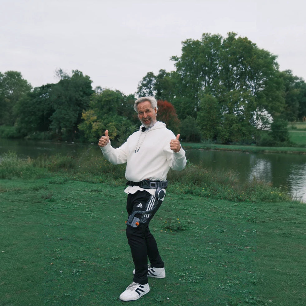 A person wearing an Ascentiz hip exoskeleton walks beside a lake and gives two thumbs up.