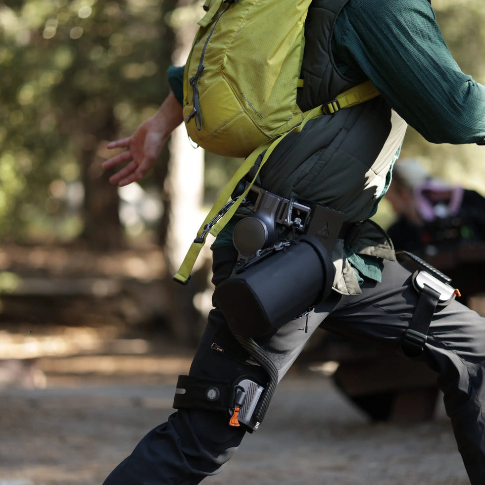 A hiker wears an Ascentiz hip exoskeleton with a backpack outdoors.