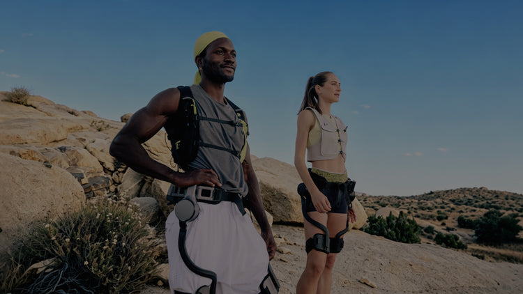 Desktop lifestyle shot of two trail runners wearing the Ascentiz H Pro hip exoskeleton in a rocky desert terrain, demonstrating lightweight wearable technology for trail running and mountain adventure.