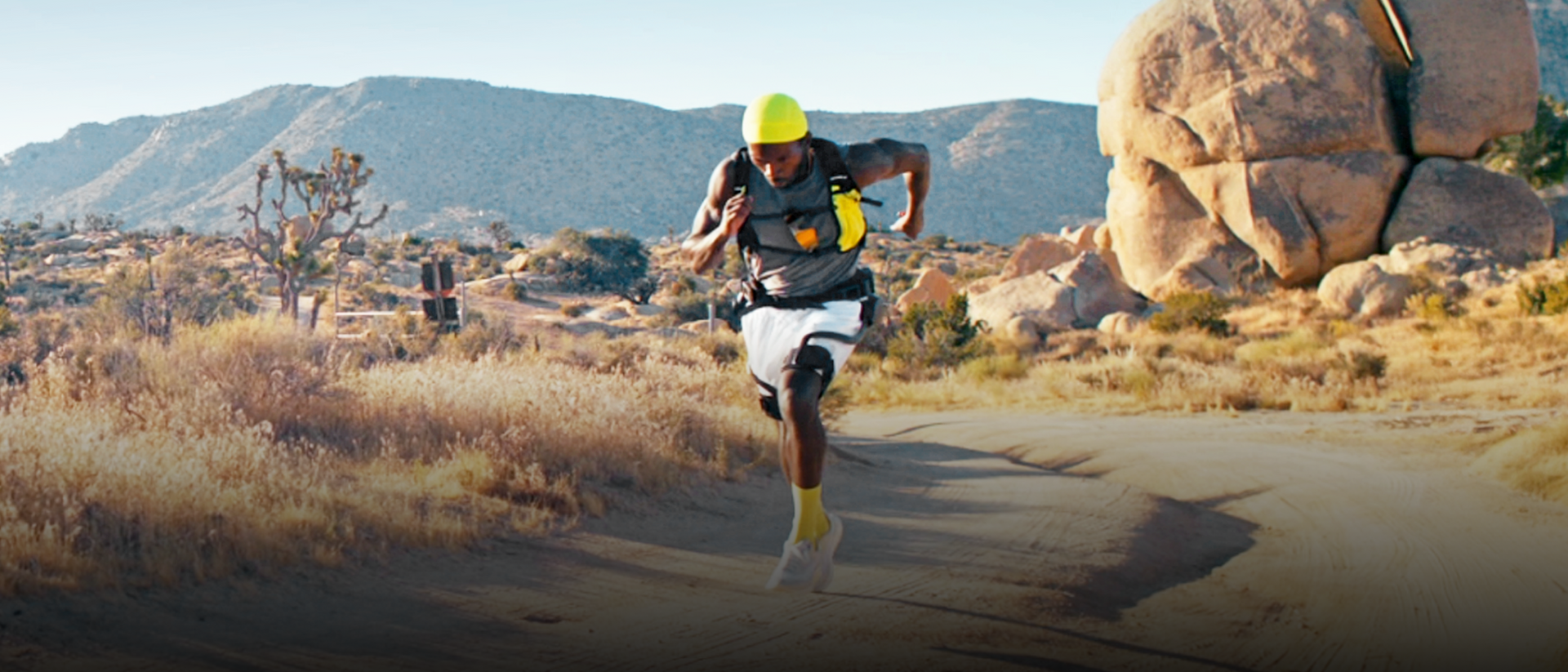 Desktop action lifestyle shot of a male trail runner wearing the Ascentiz H Pro hip exoskeleton and a yellow durag, running with speed on a rocky desert trail, showcasing technology for endurance.