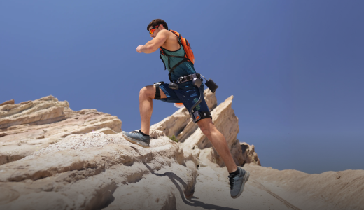 Desktop lifestyle image of a trail runner wearing the Ascentiz modular hip exoskeleton and an orange backpack while climbing a steep, rocky desert trail, showcasing thigh muscle fatigue reduction and uphill kinetic assistance.