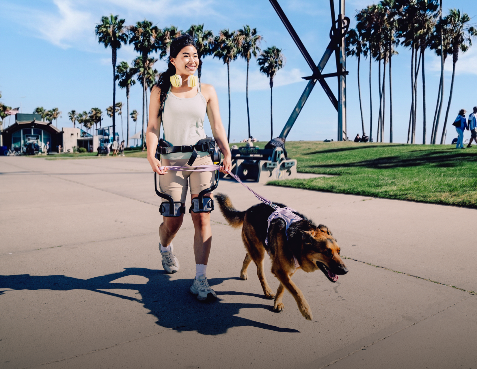 Desktop lifestyle shot of a smiling woman walking a dog on a sunny paved path while wearing the Ascentiz H Pro hip exoskeleton, showcasing daily mobility assistance and active outdoor leisure.