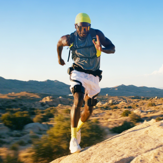 Dynamic lifestyle image of a male trail runner wearing the Ascentiz hip exoskeleton while running along a rocky desert trail under a blue sky, showcasing kinetic assistance and thigh muscle fatigue reduction for extreme outdoor sports.