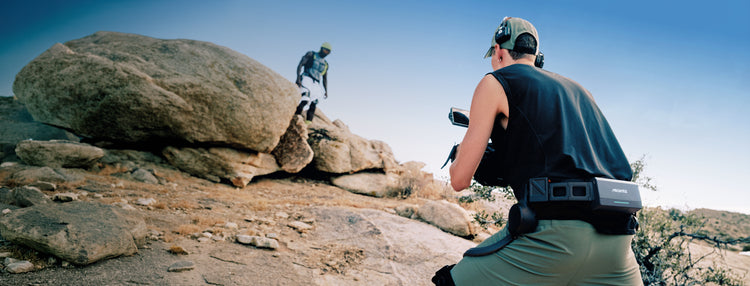 A filmmaker capturing a runner in a rugged outdoor landscape, featuring the Ascentiz hip exoskeleton which provides stable support and mobility for professional outdoor content creation.