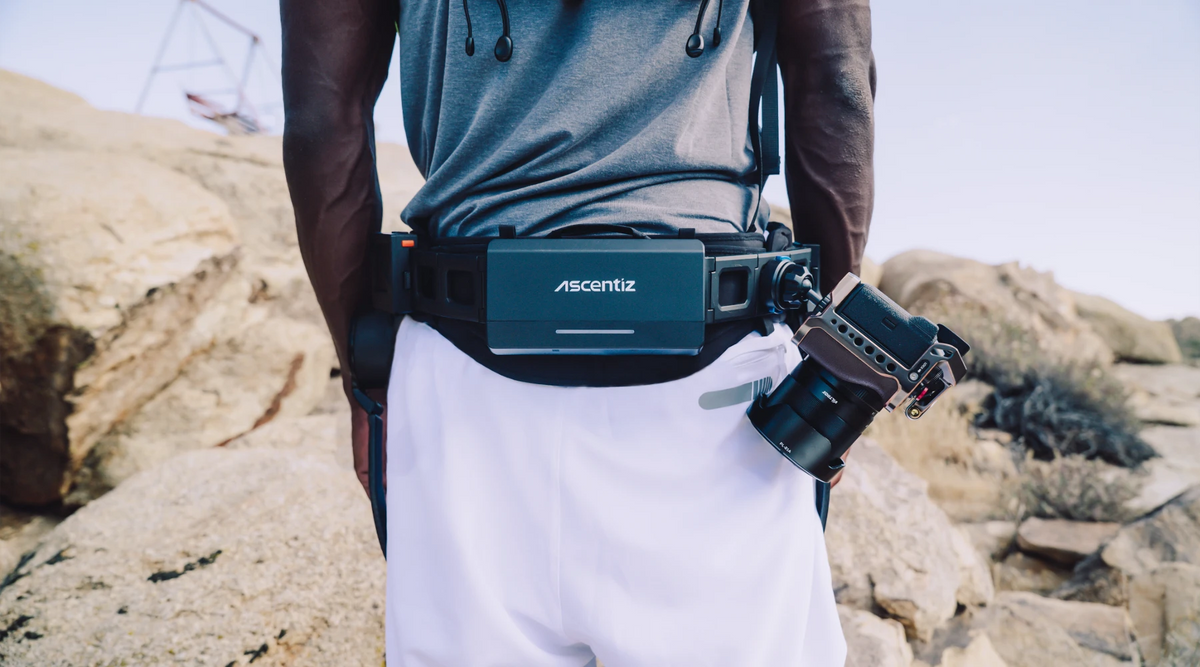 Close-up shot of a professional camera securely mounted to the Ascentiz modular hip exoskeleton belt using a dedicated attachment mount. This wearable robotic ecosystem allows wilderness photographers to maintain rapid gear access while ensuring stability and weight distribution during demanding outdoor field assignments.