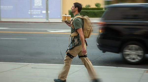 Blog lifestyle image of a delivery worker wearing the Ascentiz hip exoskeleton on a city street, carrying a heavy backpack and drinks, showcasing workplace safety and occupational load-bearing assistance.