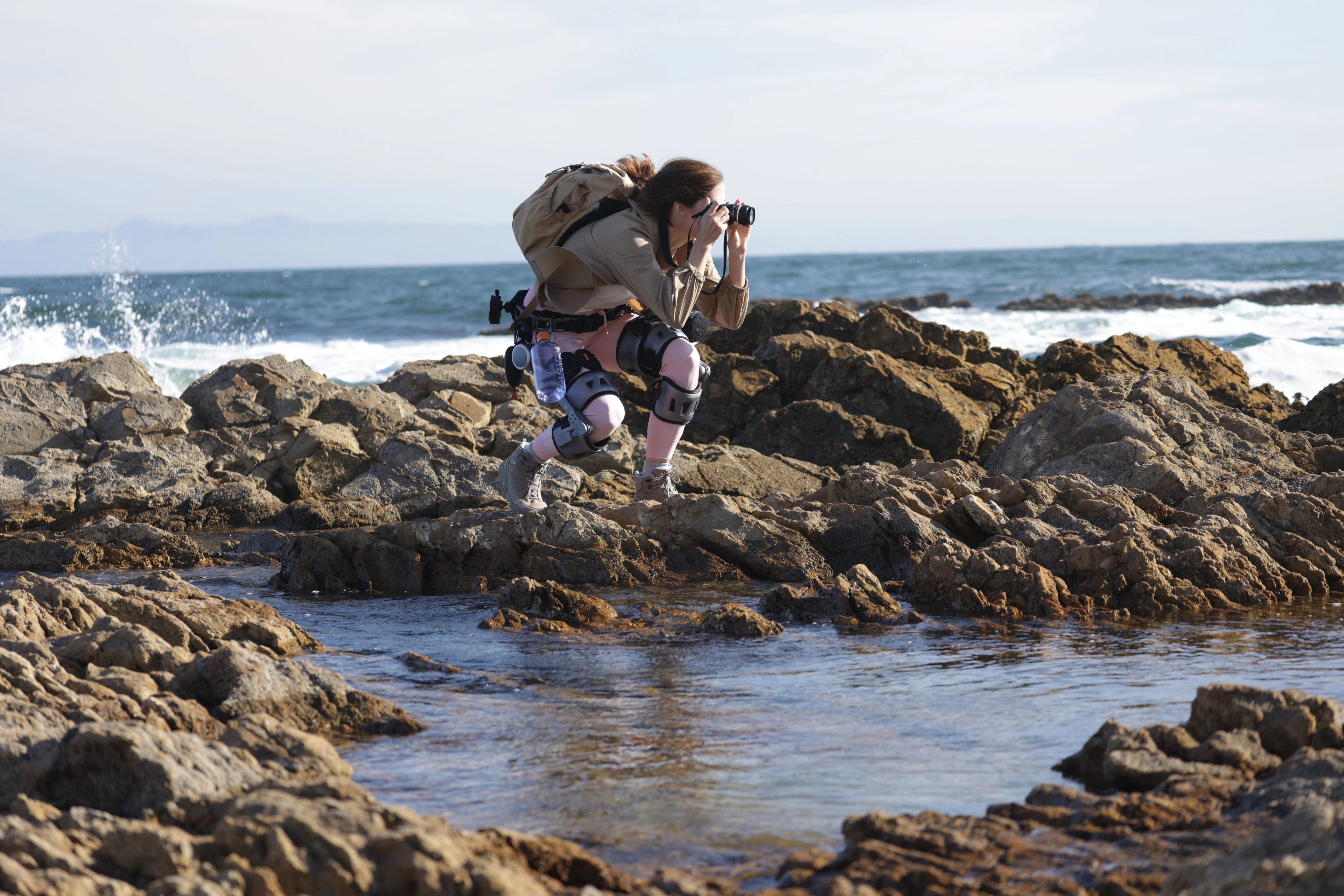 Woman wearing Ascentiz K-module exoskeleton kneeling by the sea taking photo of sea view