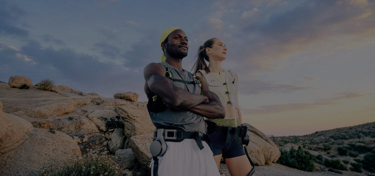 Lifestyle action shot of two hikers in a rocky desert landscape at sunset, both wearing Ascentiz modular hip exoskeletons. This image highlights the social experience of outdoor adventure, demonstrating how wearable robotic technology enhances endurance and mobility for hiking partners.