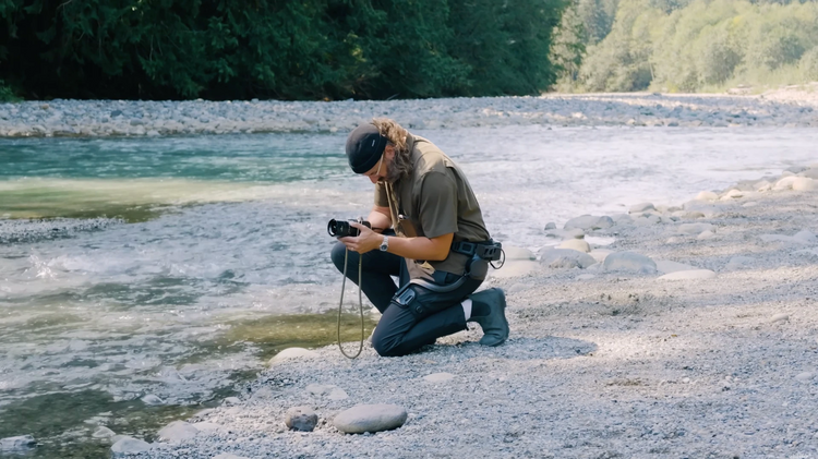 A person kneels on a rocky riverbank beside a flowing stream, holding a camera.