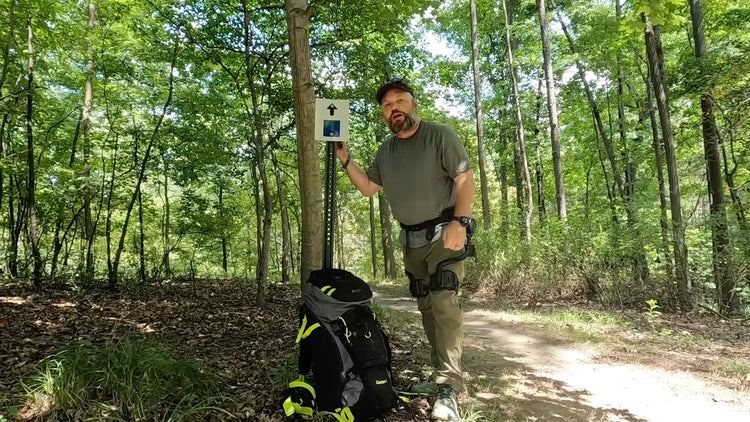 A man stands on a forest trail next to a trail sign and a black hiking backpack. He is going to make a backpacking trip through the forest with the assistance of Ascentiz robotic legs.