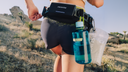 Ascentiz Exo-Belt worn by a runner outdoors, featuring rear attachment mounts with a blue Nalgene water bottle and a clear accessory pouch.