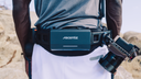 A person outdoors wearing a black waist belt with a dark teal Ascentiz device mounted on the front, alongside a camera rig.