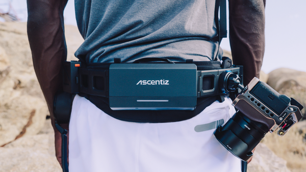 A person outdoors wearing a black waist belt with a dark teal Ascentiz device mounted on the front, alongside a camera rig.