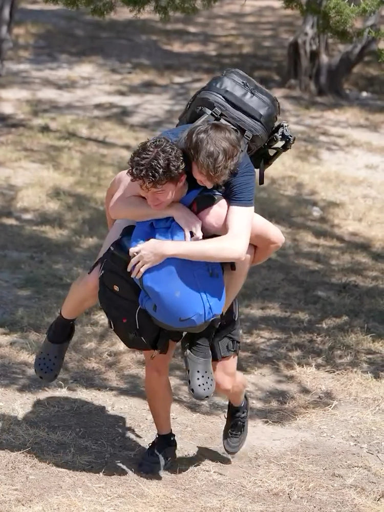 Two hikers outdoors on a trail, with one person carrying the other on their back along with three backy backpacks, while wearing the Ascentiz hip support device.